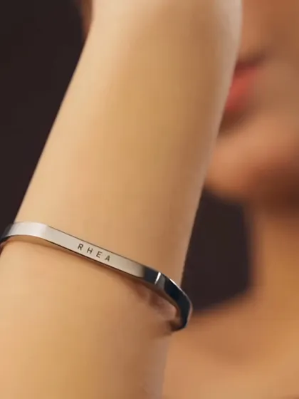 A close-up on a woman's arm, showing the delicate and clean engraving of the name 'RHEA' on a silver cuff. The minimalist design makes it perfect for daily wear.