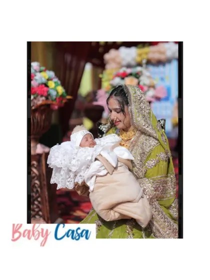 A mother holds her newborn during a traditional ceremony. The baby is dressed in a comfortable and elegant white outfit, kept cozy in a personalized wrap, perfect for family gatherings.