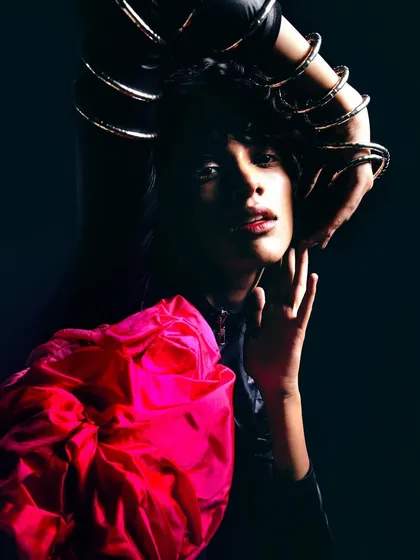 A close-up portrait with dramatic lighting. The model's hands, adorned with gold bangles, frame her face, creating an intimate and powerful composition.