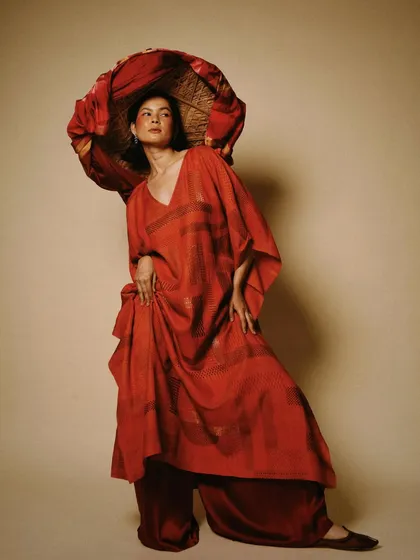 A dramatic, high-fashion pose showcasing a flowing red kaftan. The use of a traditional hat as a prop adds an artistic and sculptural element to the image.