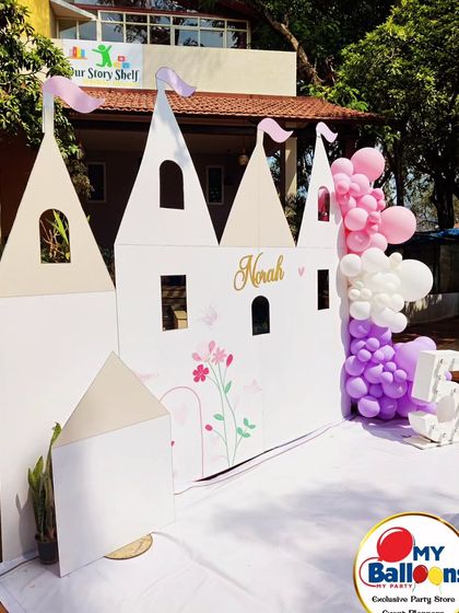 A life-sized castle backdrop for a fifth birthday. This simple yet elegant setup features a large white castle cutout, a single dessert plinth, and a purple and pink balloon garland.