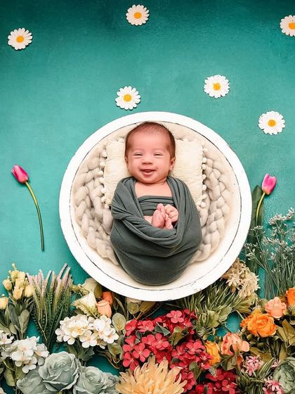A smiling newborn in a basket against a teal backdrop, with a garden of flowers at the base. A happy, awake baby makes for a precious and unique shot.