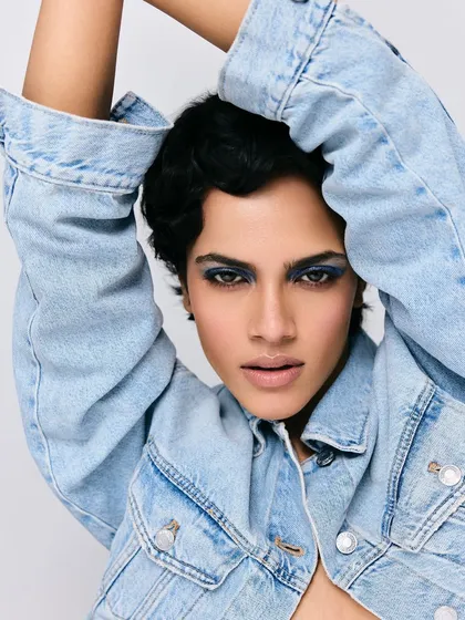 A striking portrait featuring a model with a pixie cut and bold blue smokey eyes. The styling is minimal, focusing on the denim jacket and the powerful, direct gaze.