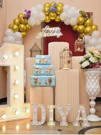 This is a popular setup for a first birthday, featuring a large light-up marquee '1'. The gold and white balloon arch, decorative birdcages, and stacked trunks create a beautiful and classic photo corner.