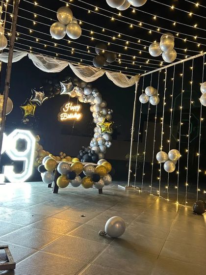 A stunning outdoor canopy decoration for a 29th birthday. The setup is draped in fairy lights and features a black, gold, and silver balloon arch with a neon sign and '29' marquee numbers.