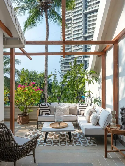 This outdoor terrace at 'Summer Haus' is an urban oasis. A wooden pergola provides partial shade for a comfortable lounge area, creating a perfect spot for relaxation amidst the lush greenery, with the Mumbai skyline in the background.