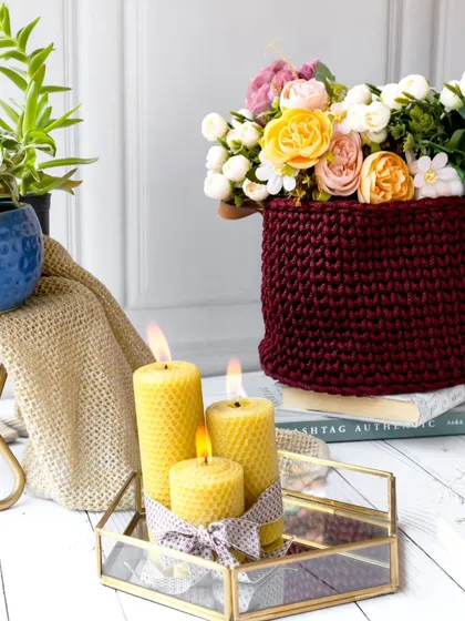 A vibrant scene combining a burgundy crochet basket full of flowers with lit beeswax candles. This composition creates a warm, inviting, and beautifully styled corner of a home.