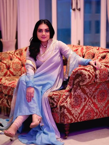 A full-length portrait showcasing the beautiful ombre effect of the light blue and lavender suit. The model sits gracefully on a vintage-style sofa.