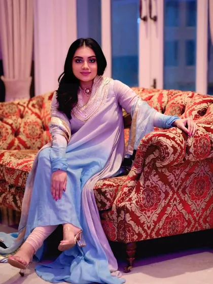 A full-length portrait showcasing the beautiful ombre effect of the light blue and lavender suit. The model sits gracefully on a vintage-style sofa.