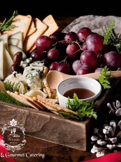 A close-up of a holiday-themed cheese box, adorned with pine cones and fresh evergreen sprigs. The selection includes various cheeses, crackers, and a dipping sauce, perfect for seasonal celebrations.