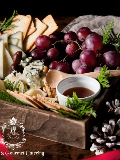 A close-up of a holiday-themed cheese box, adorned with pine cones and fresh evergreen sprigs. The selection includes various cheeses, crackers, and a dipping sauce, perfect for seasonal celebrations.