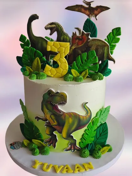 A tall dinosaur cake with realistic-looking toppers and lush green foliage. The design creates a dramatic and impressive centerpiece for a 3rd birthday party.