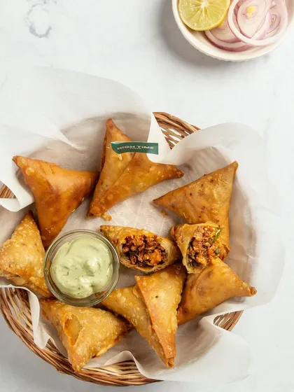 This top-down shot of plant-based keema samosas is beautifully styled in a rustic basket. The broken-open samosa reveals the hearty filling inside, showing customers exactly what they're getting.