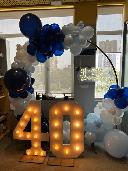 A 40th birthday celebration in an office space, featuring a bold blue and white balloon arch, a "Happy Birthday" neon sign, and large light-up marquee numbers. I can bring the party right to your workplace.