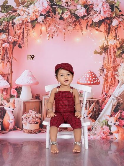 A baby boy sits on a chair in a beautiful pink-toned woodland scene. The floral arch, teepee, and deer figures make this a truly magical and enchanting portrait.