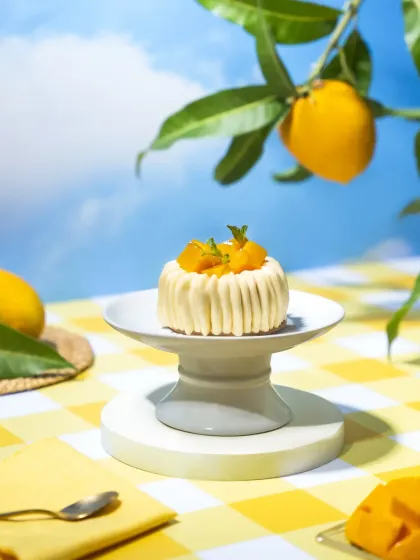 A whimsical mango mousse dessert on a pedestal, set against a dreamy blue sky backdrop with a mango branch. This conceptual shot creates a surreal summer picnic vibe, perfect for a seasonal menu.