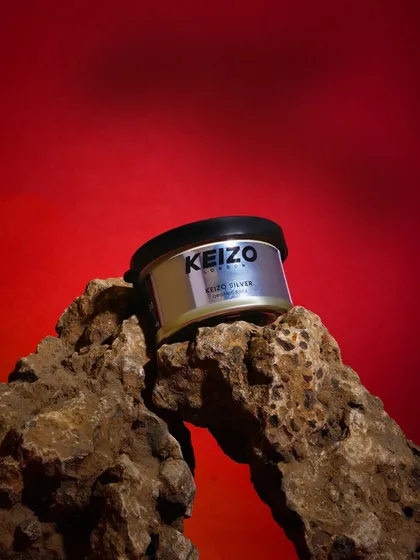 A product shot of a silver hair wax tin placed on a rough, natural rock. The contrast between the modern product and the organic element against a bold red background is striking.
