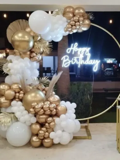 A stunning outdoor birthday setup for a night party. The large golden hoop with a neon sign is decorated with a lavish arrangement of white and gold balloons.