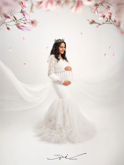 A royal portrait in white. Wearing a delicate crown and a beautiful white gown, the mother-to-be is framed by falling cherry blossoms, creating a magical and regal scene.