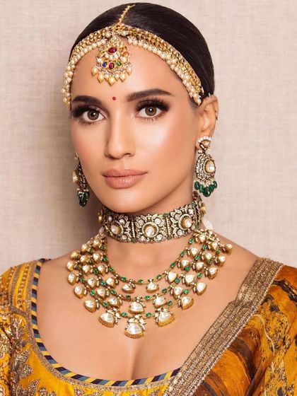 A close-up of a bride in a mustard yellow outfit. The makeup is warm and glowing, with a classic winged liner and a matha patti that frames the face beautifully.