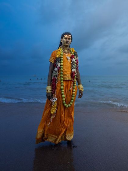 A devotee stands at the edge of the sea in Tiruchendur, the twilight sky creating a dramatic backdrop for his traditional attire and garlands.