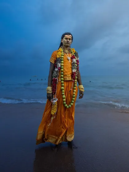 A devotee stands at the edge of the sea in Tiruchendur, the twilight sky creating a dramatic backdrop for his traditional attire and garlands.