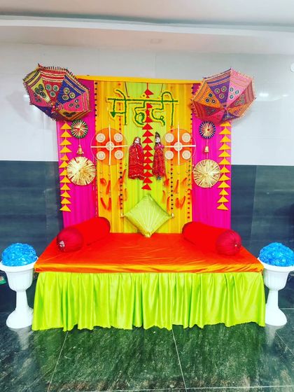 A colourful Mehendi seating area with a bright orange divan, set against a backdrop of pink and yellow drapes and decorative umbrellas.