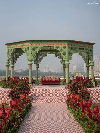 The wedding mandap viewed down the aisle, which is lined with vibrant red flowers. The design perfectly frames the couple's seating area against the city backdrop.