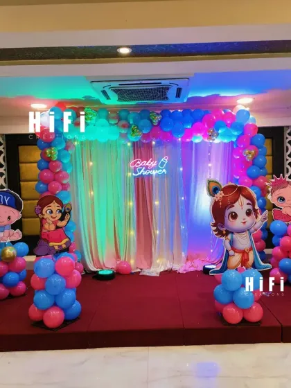 A baby shower stage with a Little Krishna theme. The decor features pink and blue balloon pillars, character cutouts, and a backdrop with elegant drapes and a neon sign.
