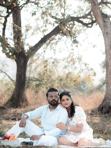 A serene, romantic picnic-themed pre-wedding shoot, with the couple sitting together under large trees in a sunlit field. This shows how we can create a beautiful, story-driven session in a natural outdoor setting.