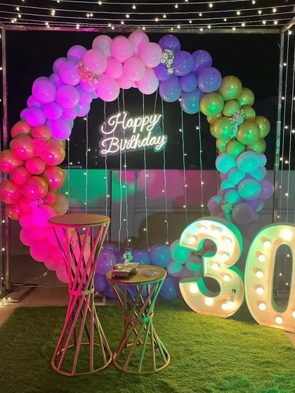 This vibrant 30th birthday celebration on a terrace features a rainbow-themed balloon arch and a 'Happy Birthday' neon sign. The setup is framed by a cube of fairy lights and includes large, illuminated '30' marquee numbers.