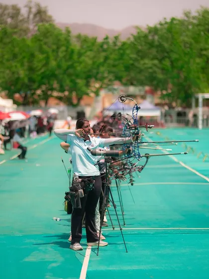 A photograph from the National Women's Archery Tournament in Ambaji, showing a line of archers taking aim. This image captures the scale of the event and the collective focus of the athletes.
