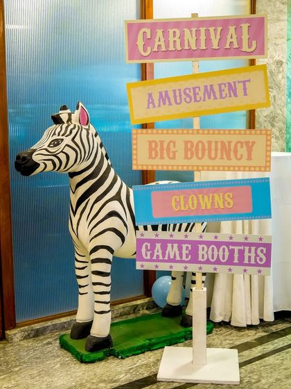 A signpost for the pastel carnival, pointing the way to the bouncy castle, clowns, and game booths, with a zebra prop standing by.