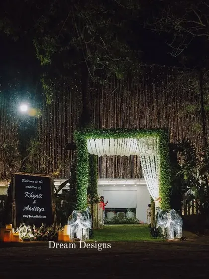 The grand entrance to a nighttime wedding. A leafy archway, a personalized welcome sign, and two decorative elephants create a memorable and traditional welcome for guests.