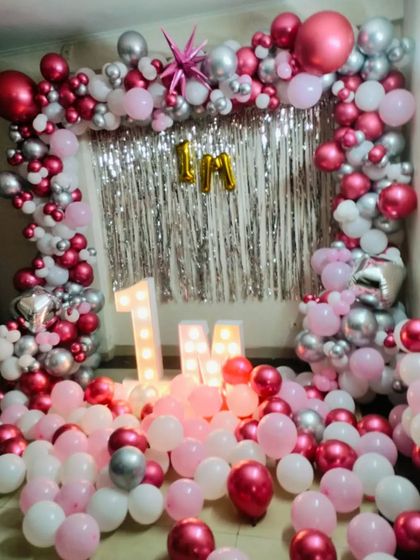 A festive room filled with pink, white, and silver balloons for a one-month celebration. The decor includes a balloon arch over a shimmer backdrop and light-up '1 M' letters.