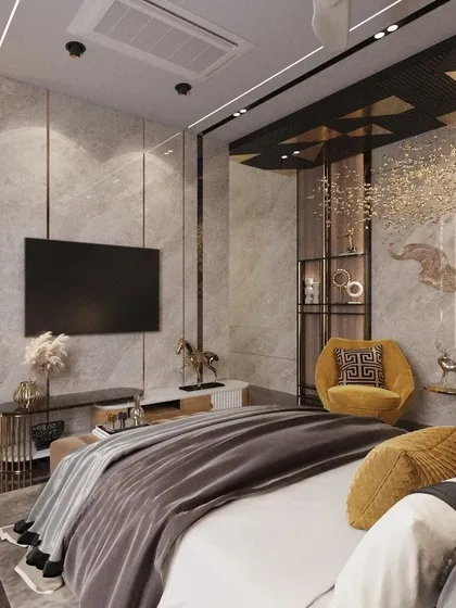 This master bedroom design combines cool grey marble with warm metallic accents for a sophisticated, modern look. A custom shelving unit and integrated TV panel create a functional yet stylish feature wall, complemented by bold yellow armchairs.