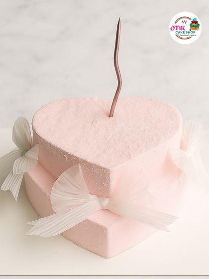 A romantic two-tiered heart-shaped cake in a soft pink velvet texture. The simple white tulle bows add a touch of grace, making it perfect for an anniversary or a romantic birthday.
