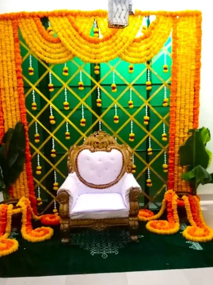 A traditional flower decoration for a Half Saree ceremony at home. The backdrop features a green cloth with a golden lattice pattern and marigold garlands.