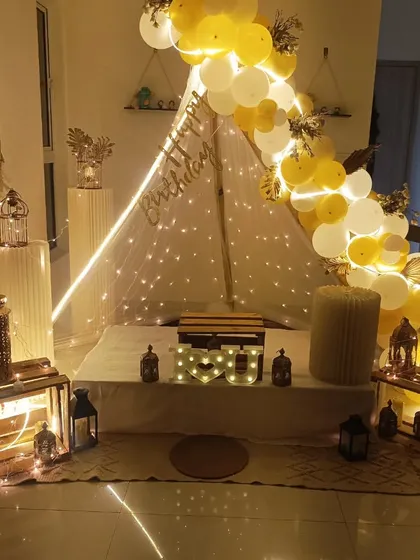 My cozy cabana tent setups are perfect for creating an intimate and memorable surprise right at home. This birthday decoration features a beautiful tent draped with fairy lights, a yellow and white balloon garland, and rustic lanterns to create a warm, magical atmosphere.