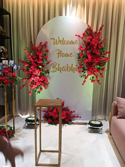 A sweet "Welcome Home Bhabhi" decoration. The white arched backdrop with gold lettering is flanked by bold red floral arrangements.