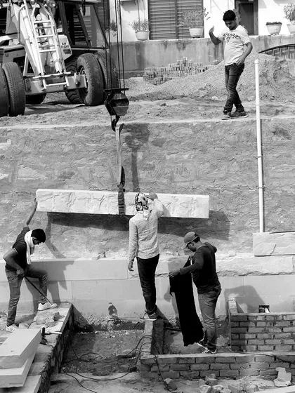 A black and white shot capturing the raw effort of construction on the House of Solid Stone. Workers guide a heavy stone lintel, lowered by a crane, into its final position, a moment of precision and teamwork.