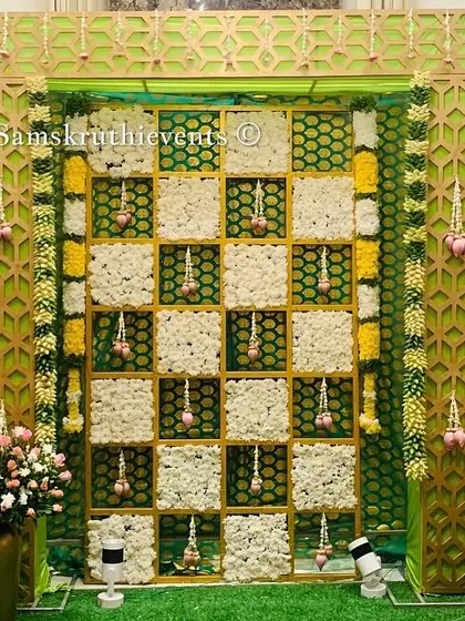 A beautiful and intricate backdrop for a traditional ceremony. The design features a checkerboard of white flowers and green panels, framed by a geometric gold border and hanging floral tassels.