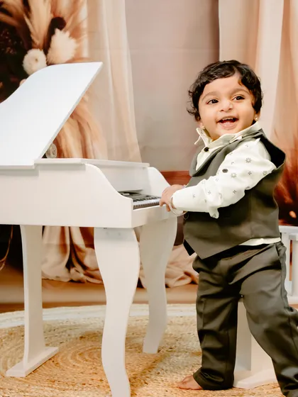 A little maestro in the making! This happy baby boy is all smiles as he stands by the miniature piano in a cozy, warm-toned studio setup.