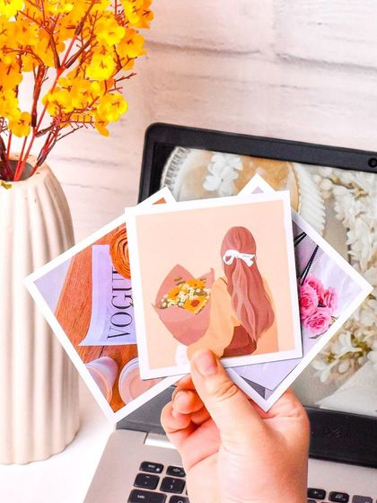 A lifestyle shot of a hand holding up several aesthetic prints in front of a laptop. This shows the product in a real-world, creative context.