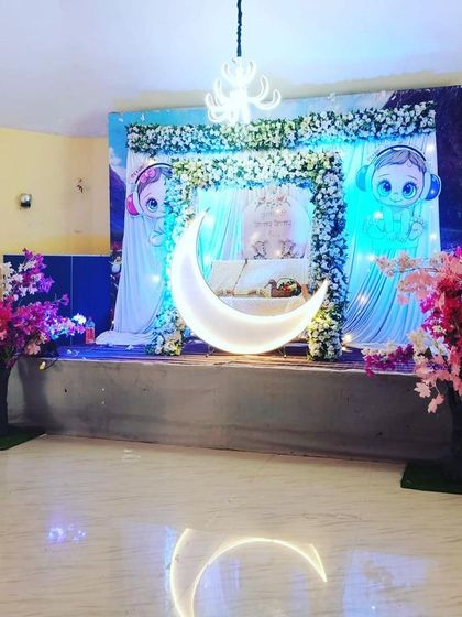 A dreamy baby shower setup with a celestial theme. The stage features a large illuminated crescent moon, character cutouts, and soft blue lighting to welcome the baby-to-be.