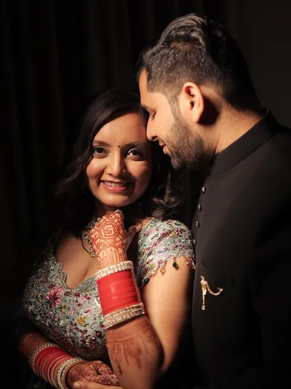 An intimate portrait from an engagement or Roka ceremony. The bride smiles at the camera while the groom looks at her adoringly. The moody lighting creates a close, personal feel, highlighting her intricate henna and red chooda bangles.