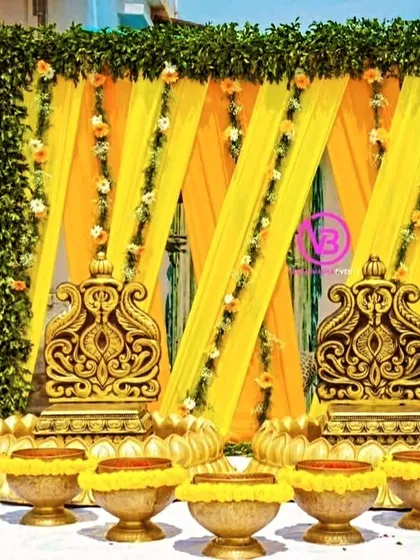 A beautiful Haldi setup with ornate golden chairs, a backdrop of yellow drapes and green foliage, and traditional brass bowls.