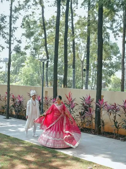A walk to remember. The bride's vibrant pink lehenga flows beautifully as she and her groom stroll through a path lined with tall, majestic trees, creating a cinematic and artistic wedding portrait.