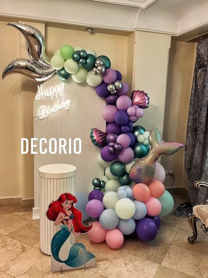 An enchanting mermaid-themed birthday decoration. This setup includes a Little Mermaid cutout, a balloon garland in ocean-inspired colors, and foil balloons shaped like mermaid tails and seashells.