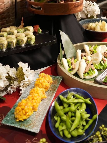 This catering spread includes a variety of textures and flavors, from steamed edamame and crispy corn fritters to soft bao buns and fresh sushi rolls.